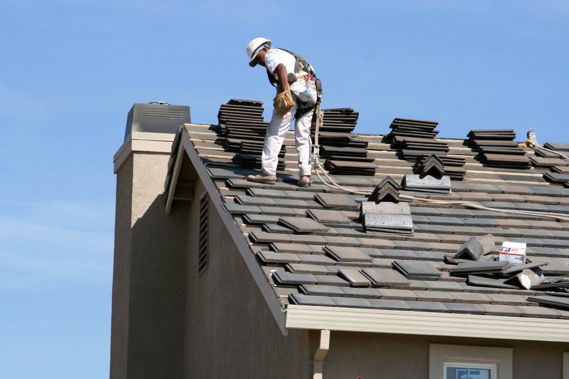 Roofer in white protective gear, working on a tiled roof against a blue sky.