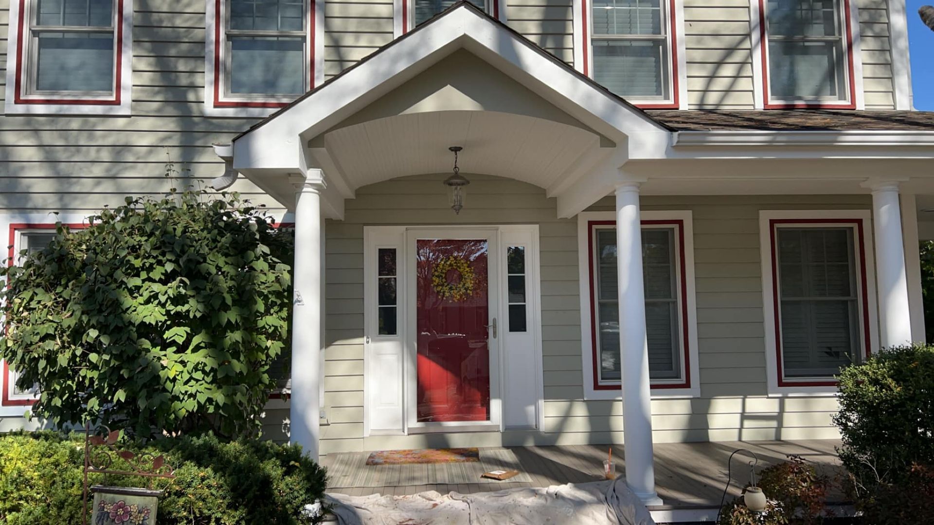 Two-story light green house with white porch, red door, and windows with red trim, sunny day.