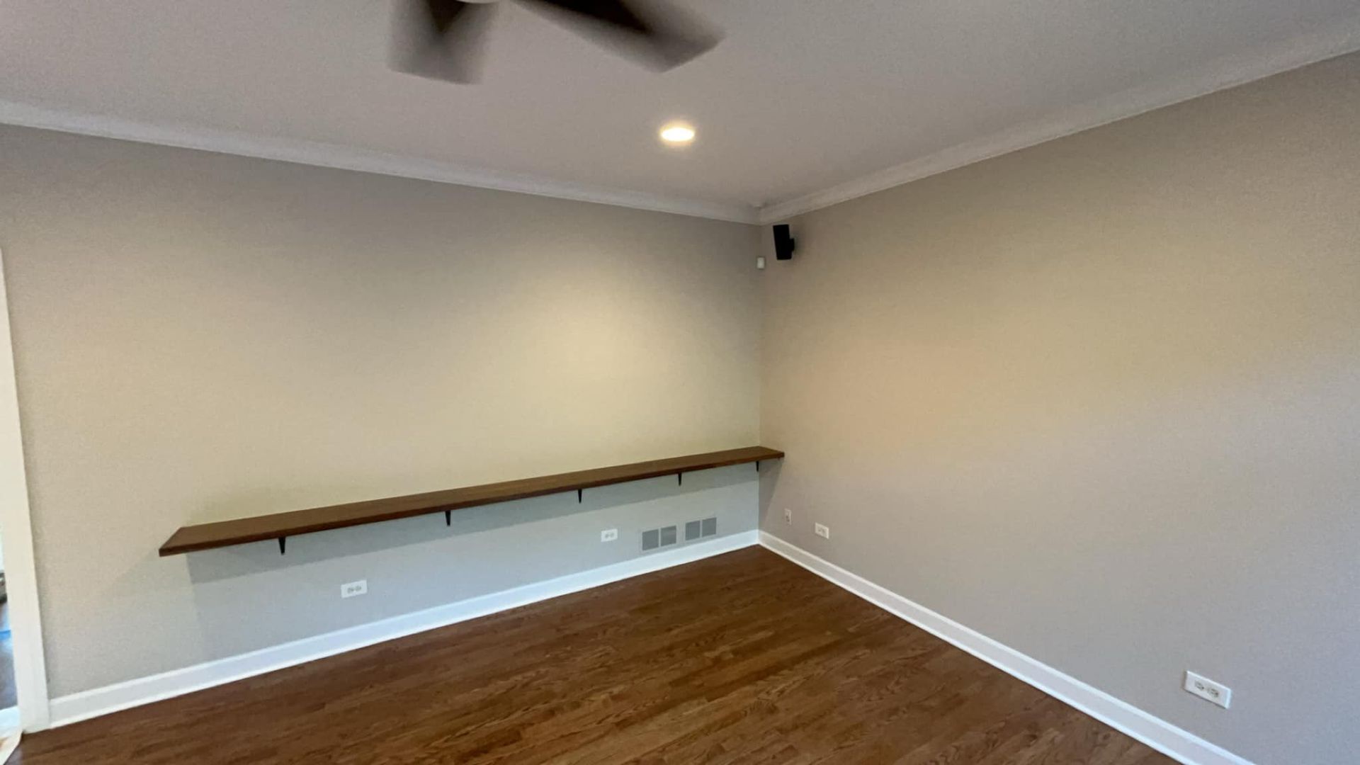A room with a long shelf on a wall, wood floor, and ceiling fan.