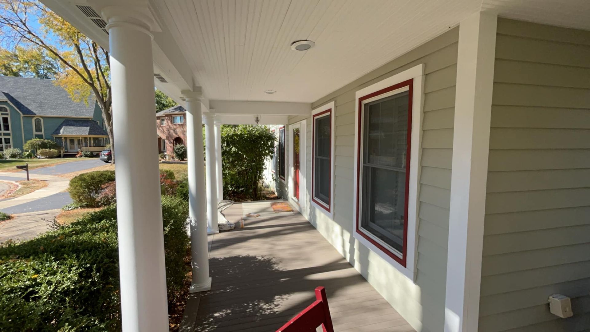 Porch with white columns and light green siding. Red window trim and a red chair.