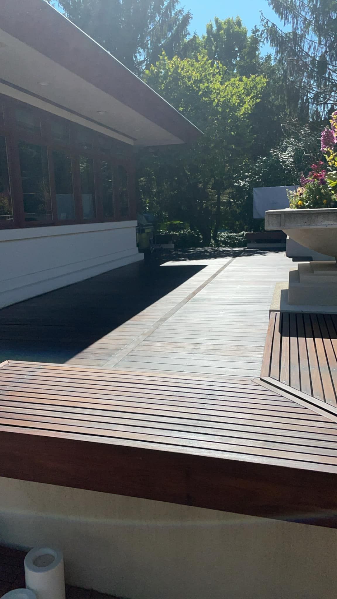 Outdoor walkway with perforated bench, white wall, trees, and a vehicle in the distance under a blue sky.