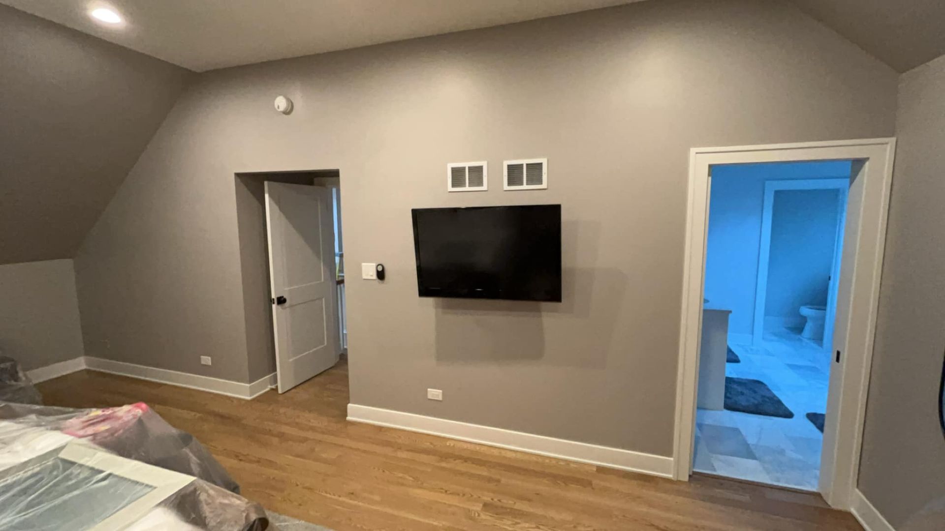 Bedroom with gray walls, hardwood floor, mounted TV, open doors to bathroom and another room.