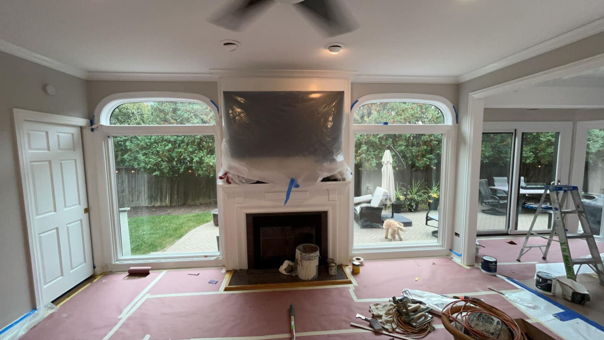 Living room with fireplace, windows, and doors; under renovation, protective covering on furnishings.