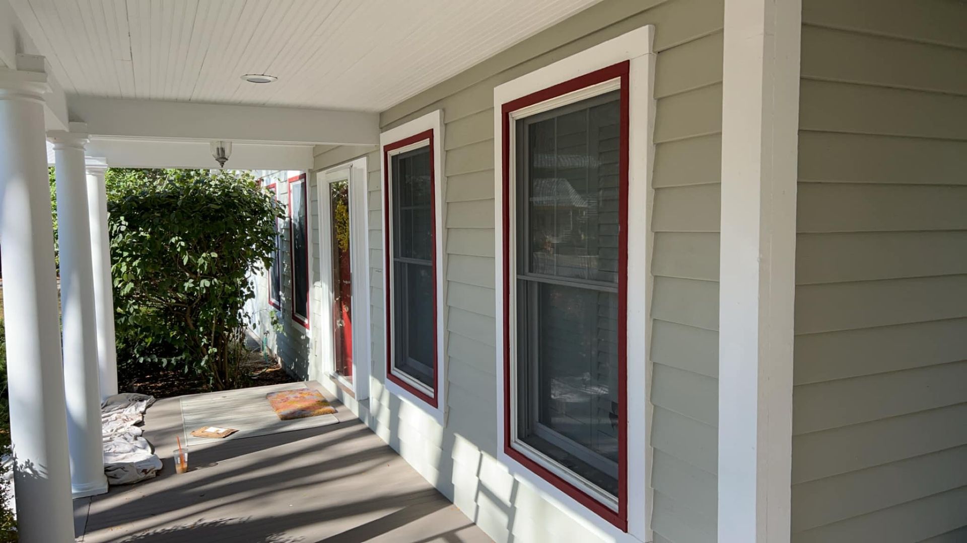 Porch with two windows framed in red, light green siding, white pillars, and bushes.