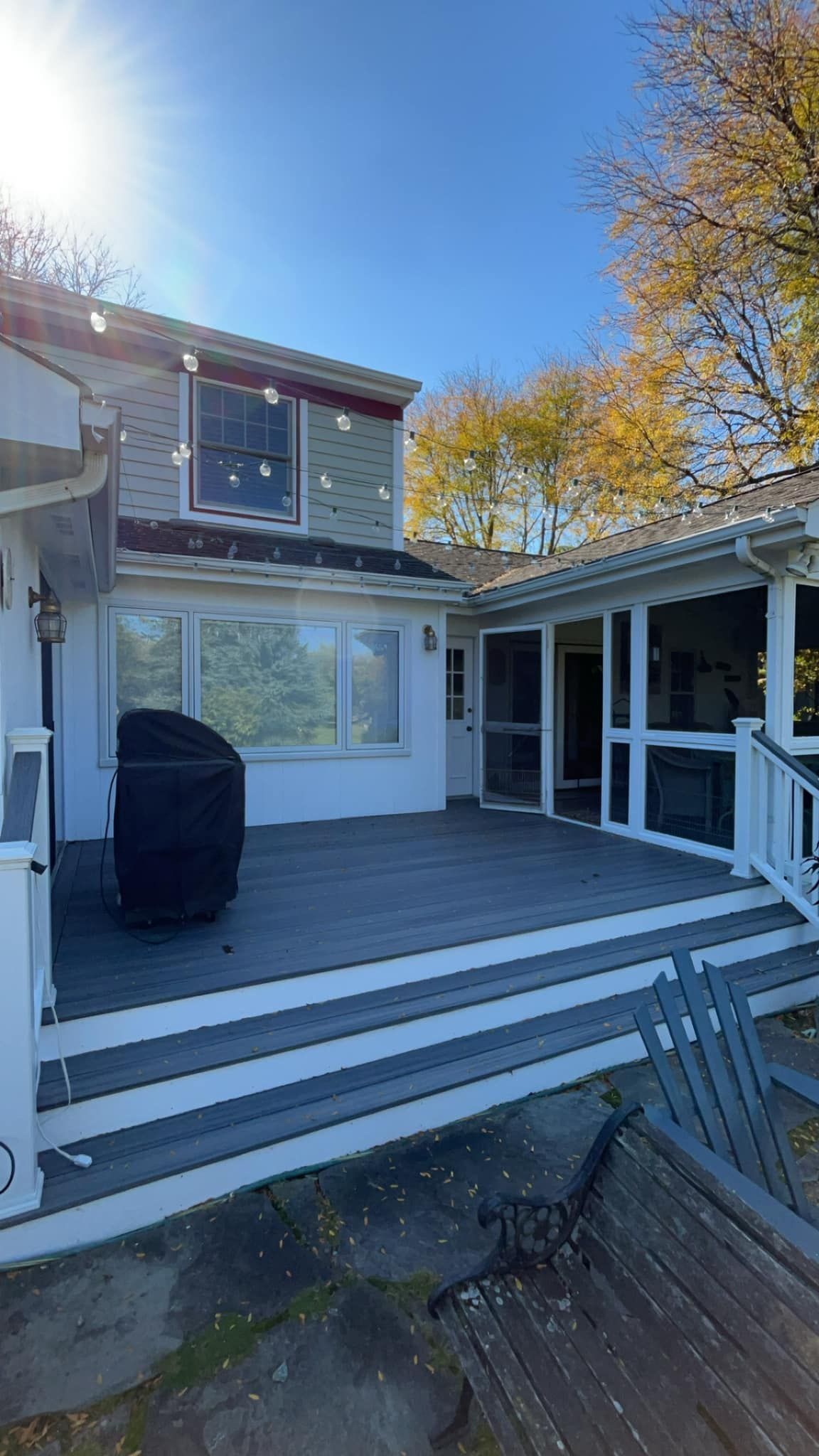 Exterior deck with gray and white railings, grill, and screened porch next to a two-story house under a blue sky.