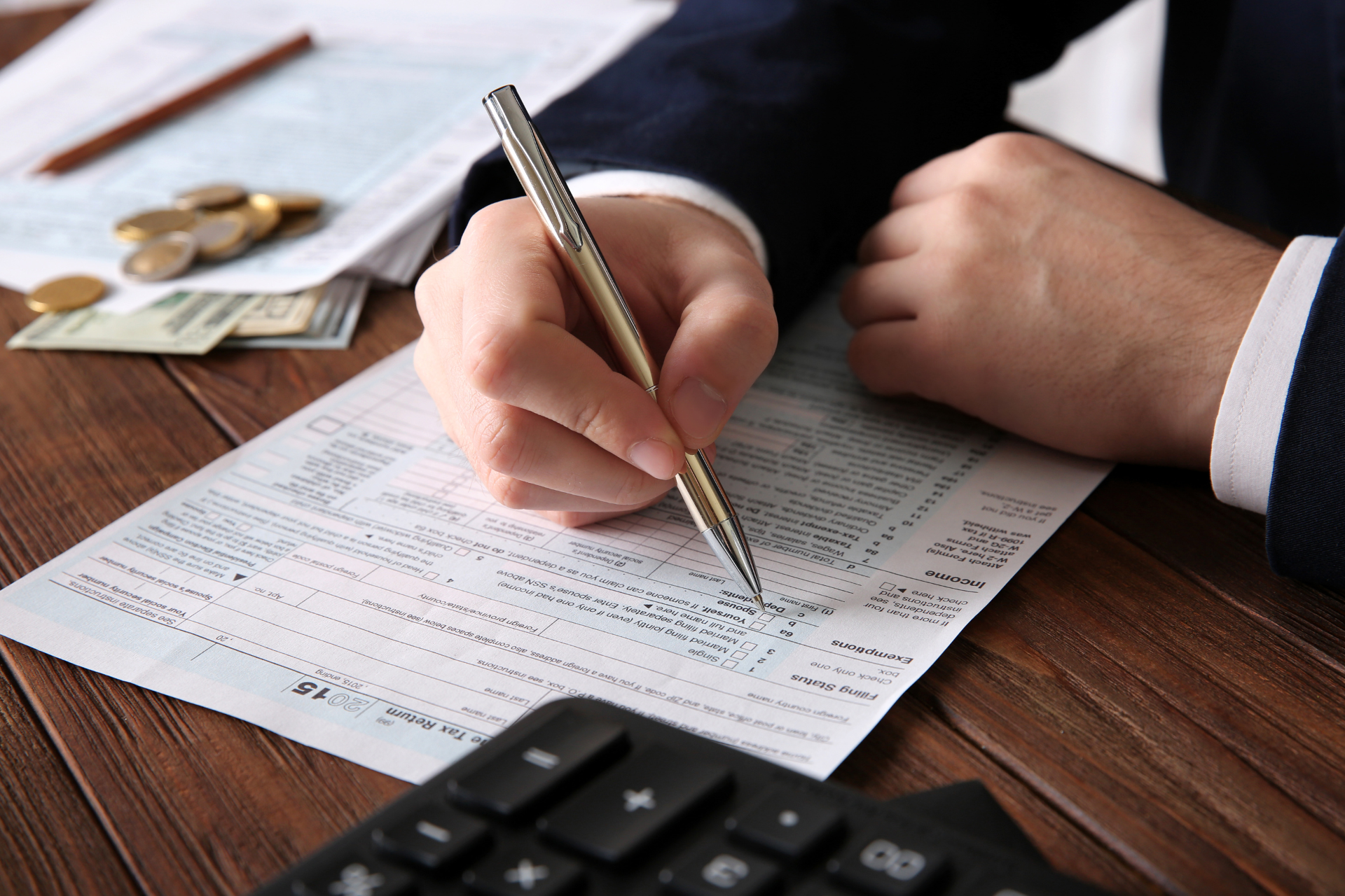Person in suit writes on a document with a pen, with money, calculator, and paperwork on a wooden desk.