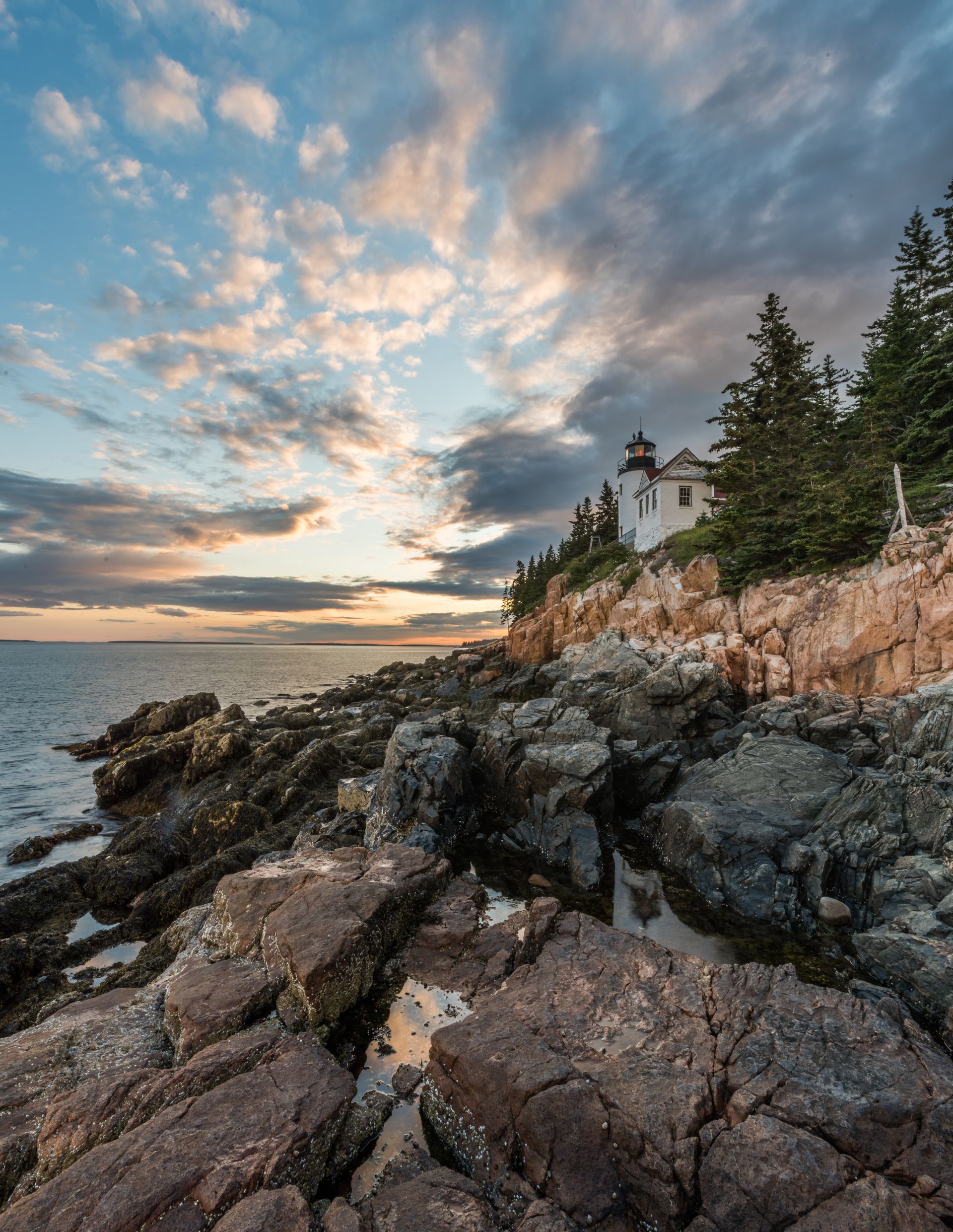 Rocky coastline with a lighthouse at sunset. Sky is cloudy with streaks of pink, orange, and blue.