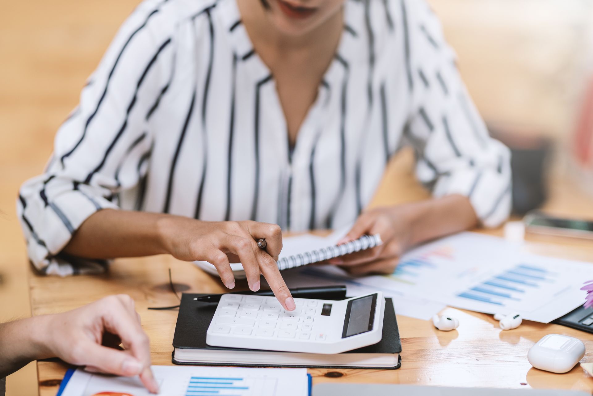 Woman using a calculator, notebook, and charts at a wooden table with another person's hand visible.