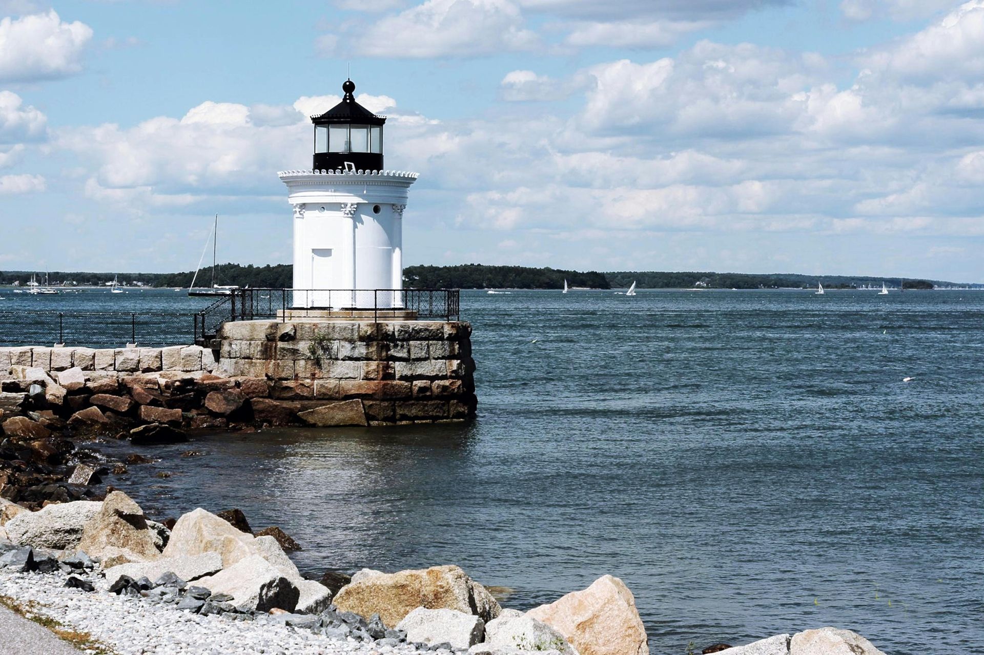White lighthouse on a stone pier, blue water, and cloudy sky.