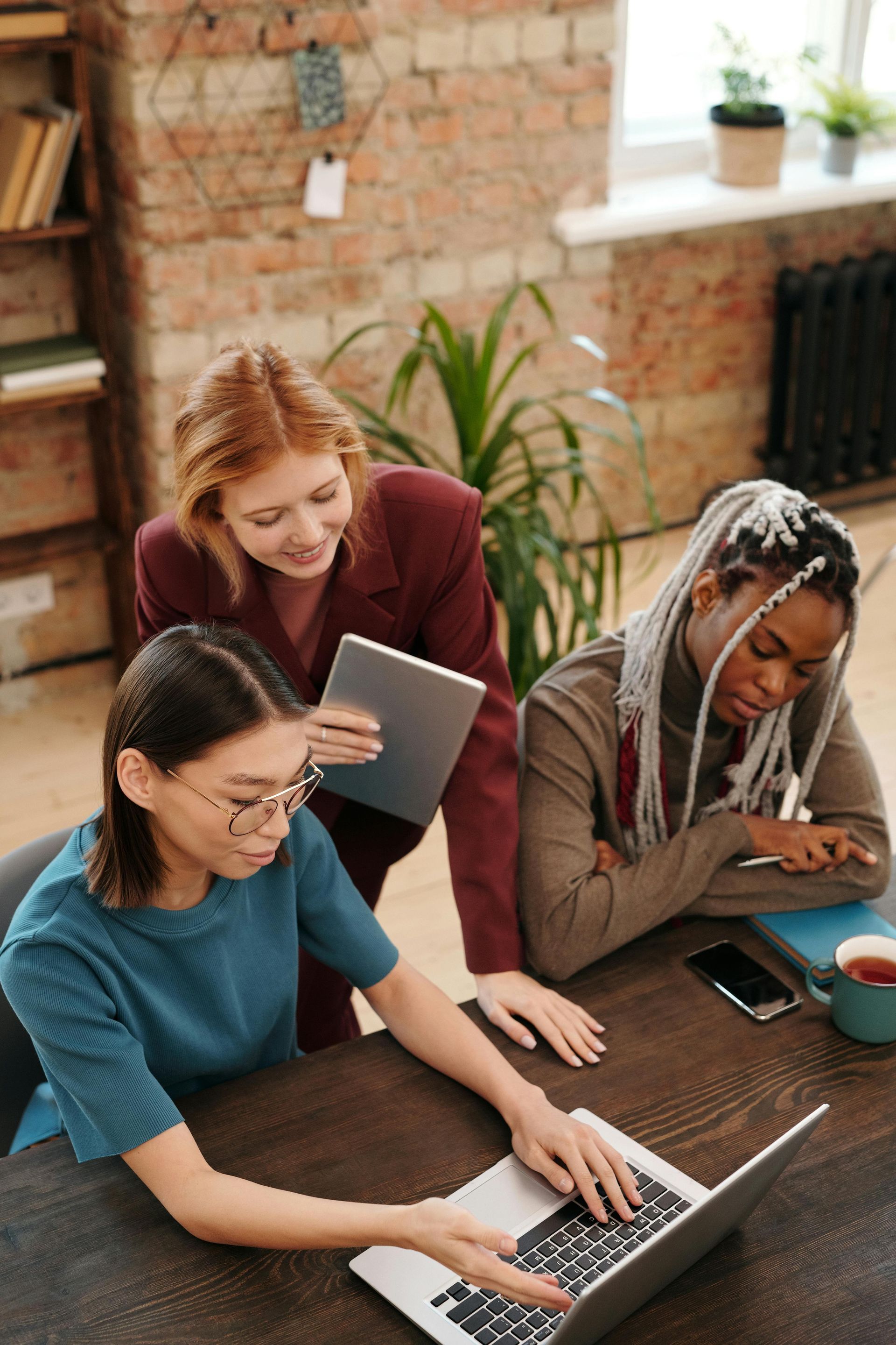 Three diverse women collaborating at a table, one using a laptop while others observe.