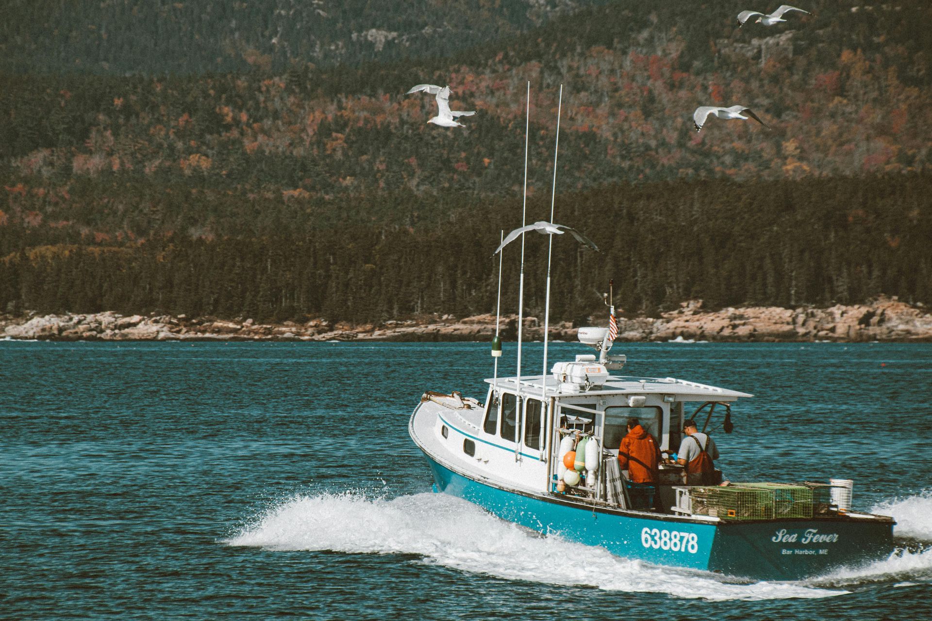 Lobster boat on blue water; two people aboard. Forested shoreline in background.