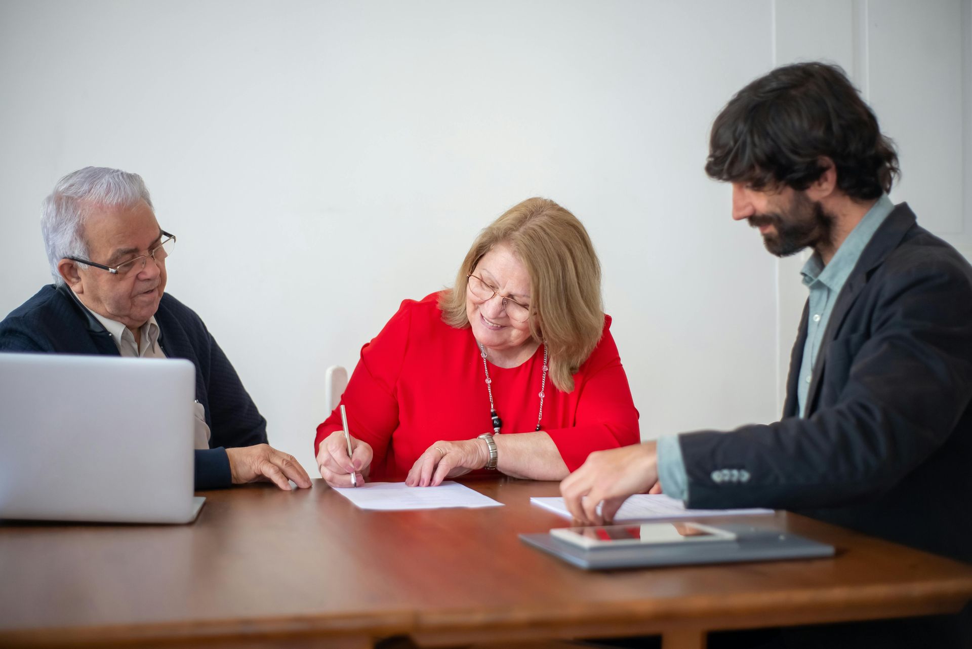 Two older adults and a younger man at a table. The woman in red signs a document.
