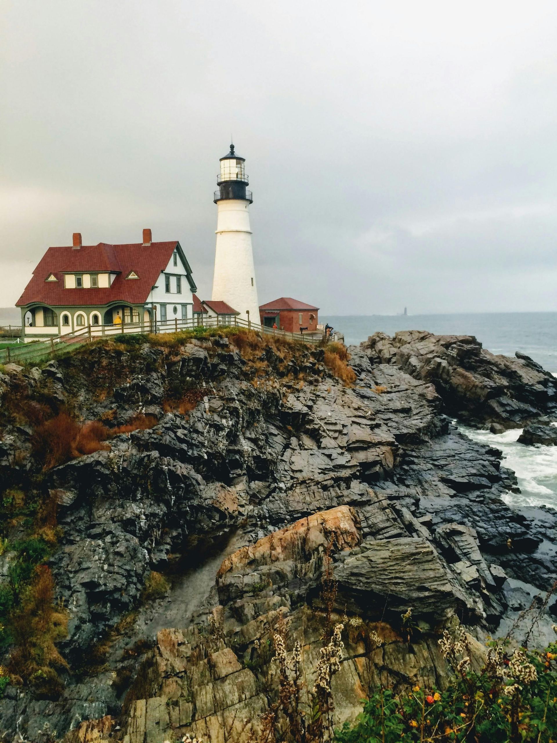 Lighthouse with red-roofed building on rocky coast under overcast sky. Ocean waves crash below.