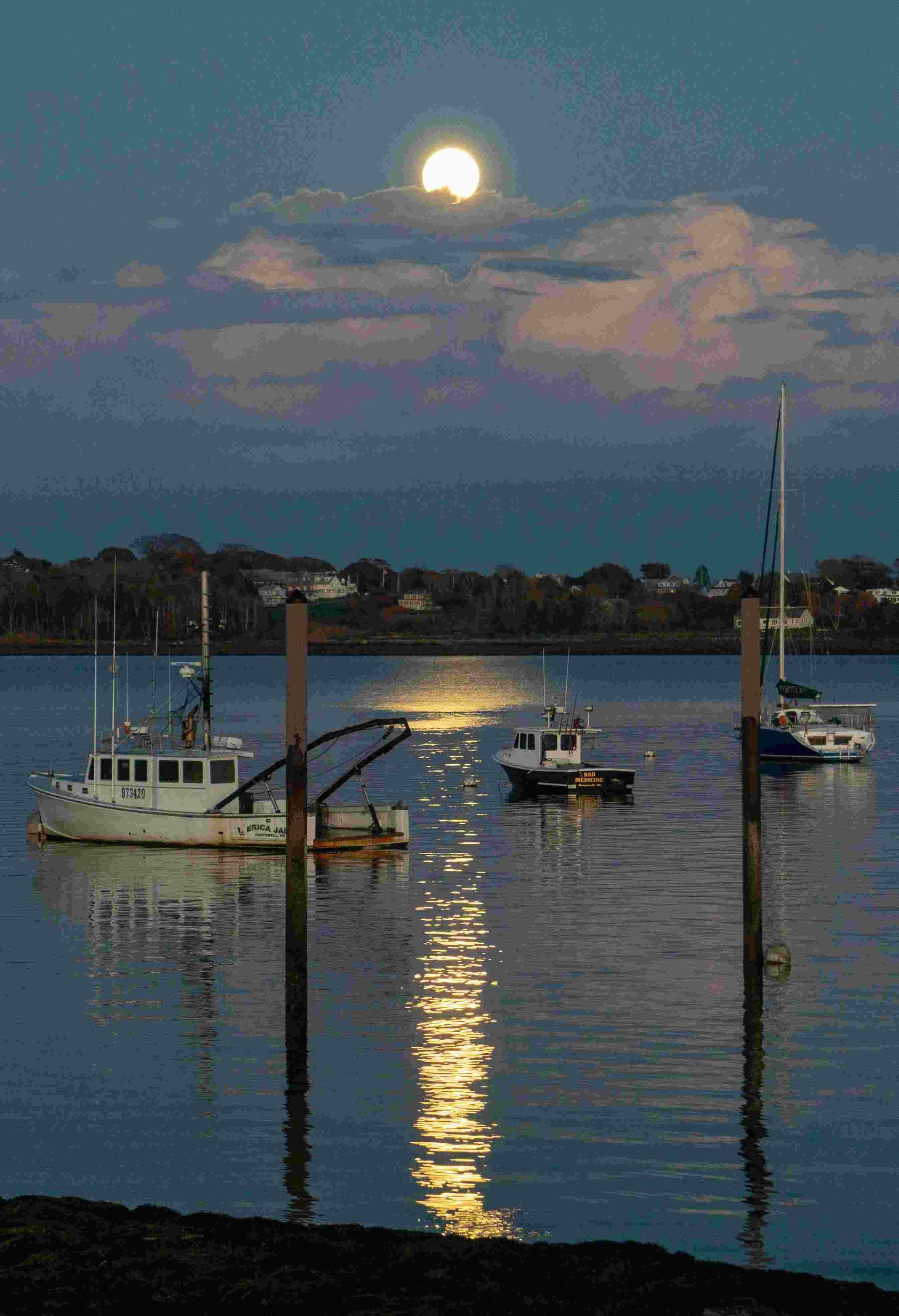Boats on calm water at dusk with a full moon, reflecting light.