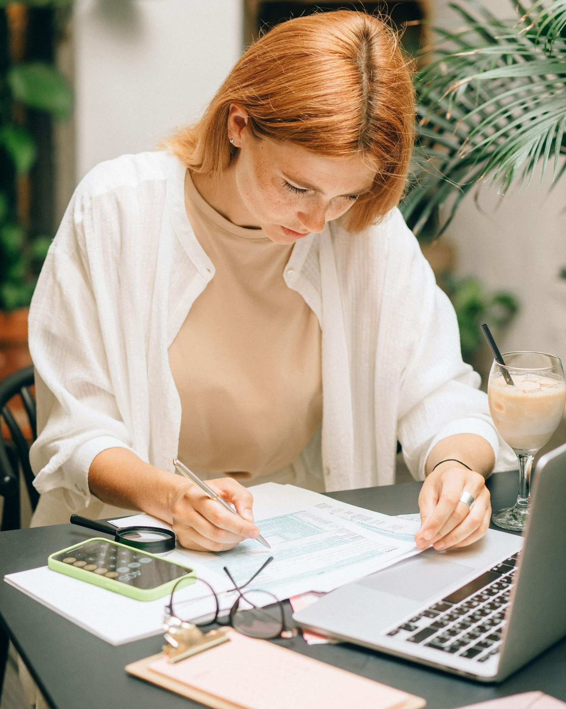 Woman with red hair, glasses, writing at table with laptop and drink.