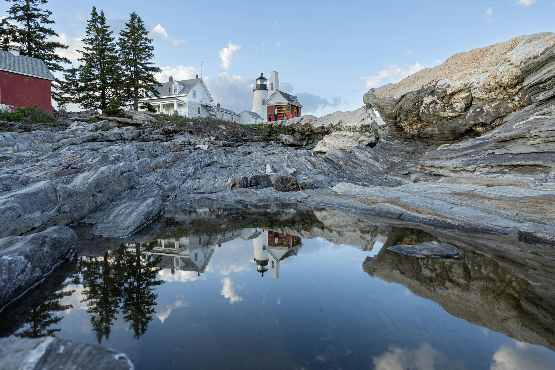 Rocky coast with buildings and a lighthouse reflected in a tide pool under a blue sky.