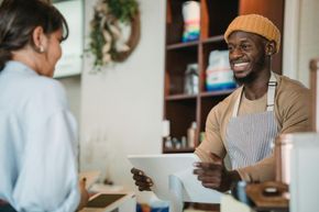 Smiling Black barista, wearing a tan knit cap and striped apron, hands a customer a tablet at a cafe counter.