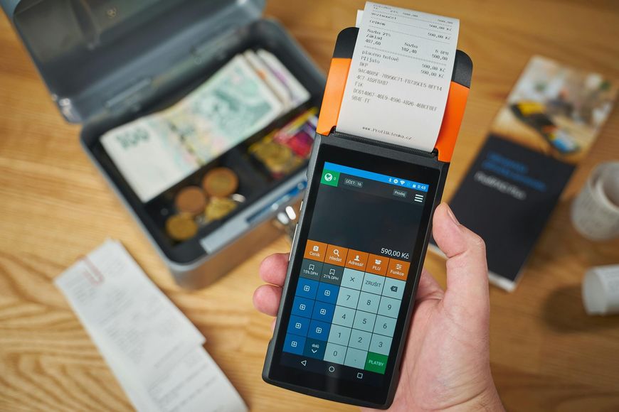 Hand holding a point-of-sale device printing a receipt, cash box open with money and coins, on a wooden surface.