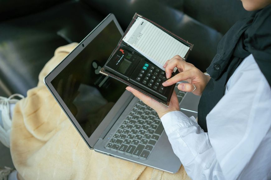 Person using a laptop, calculator, and notepad. They are seated on a couch.