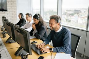 Four people with headsets work at computers in an office, smiling and typing.