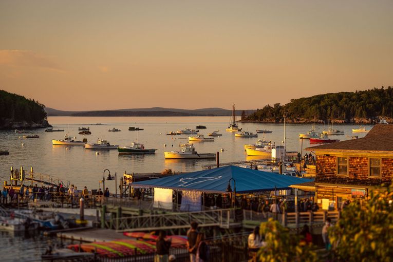 Boats in harbor at dusk with a blue-roofed restaurant and people on a pier.