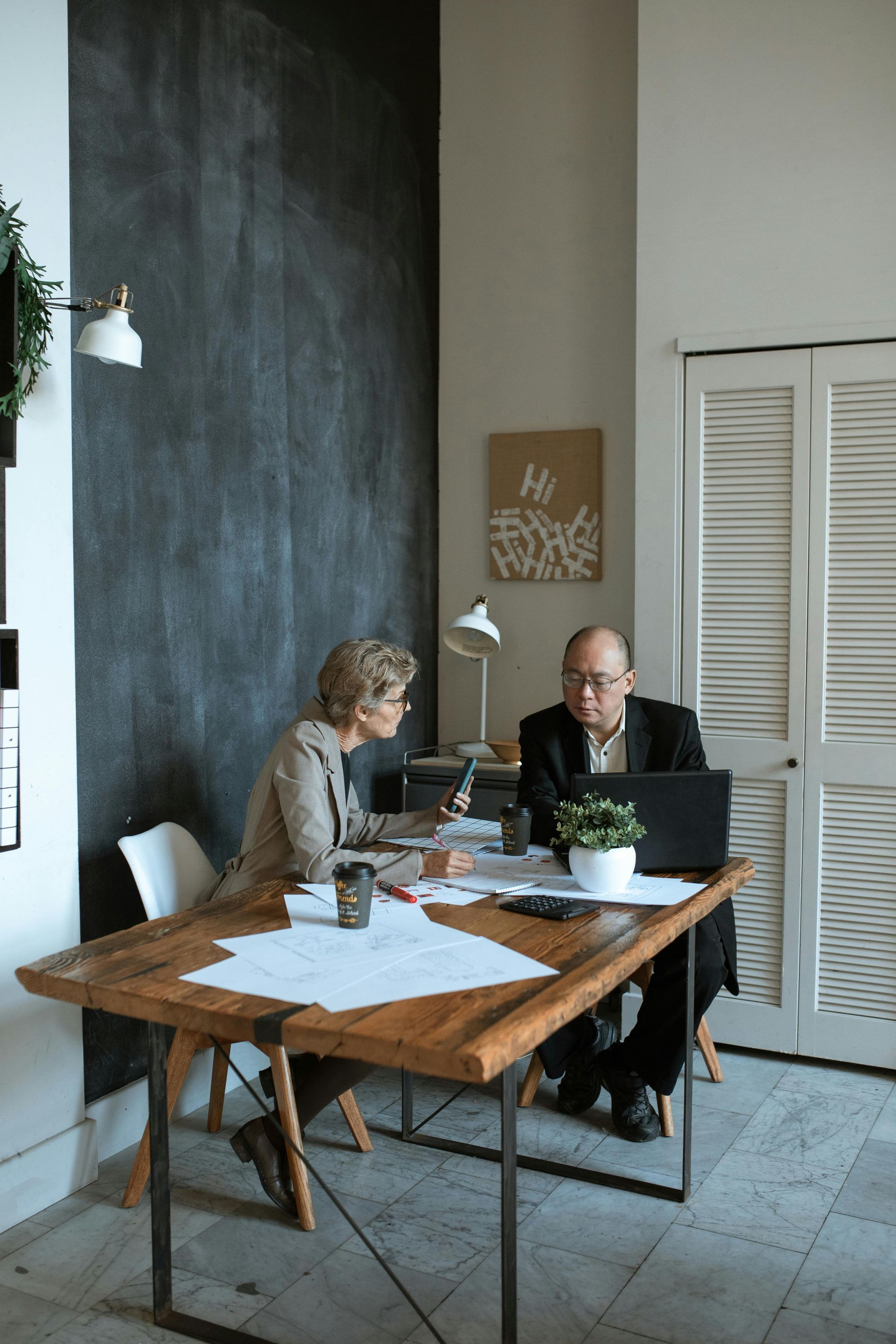 Two people in an office setting, reviewing documents and a laptop at a wooden desk.