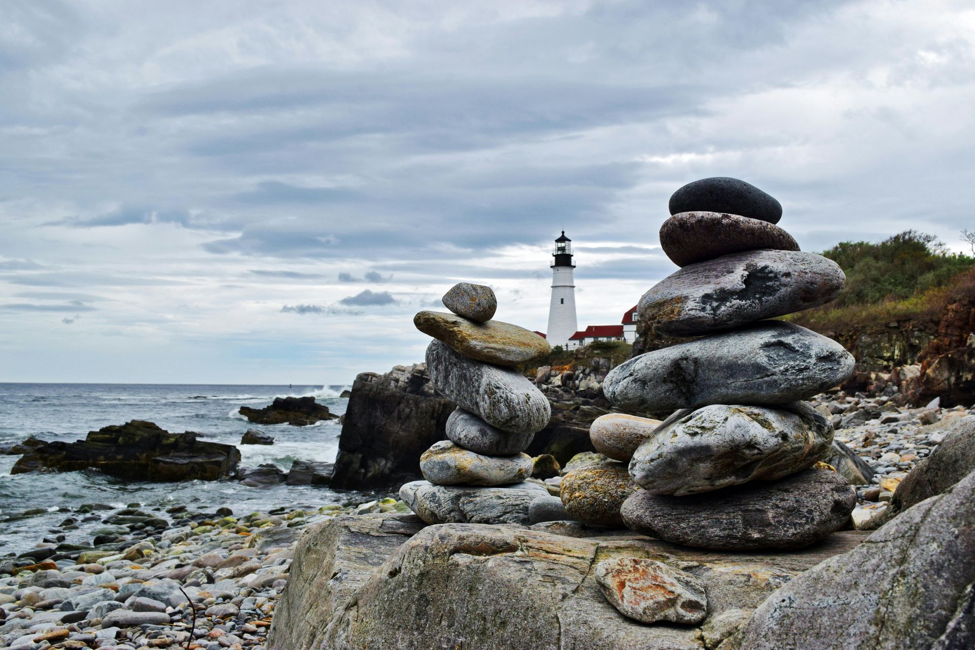 Stone cairns on a rocky coast, with a lighthouse in the background under a cloudy sky.