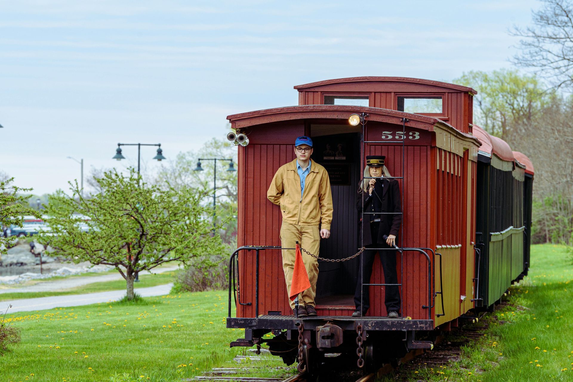 Two people in train conductor attire standing on a red caboose, in a grassy area.