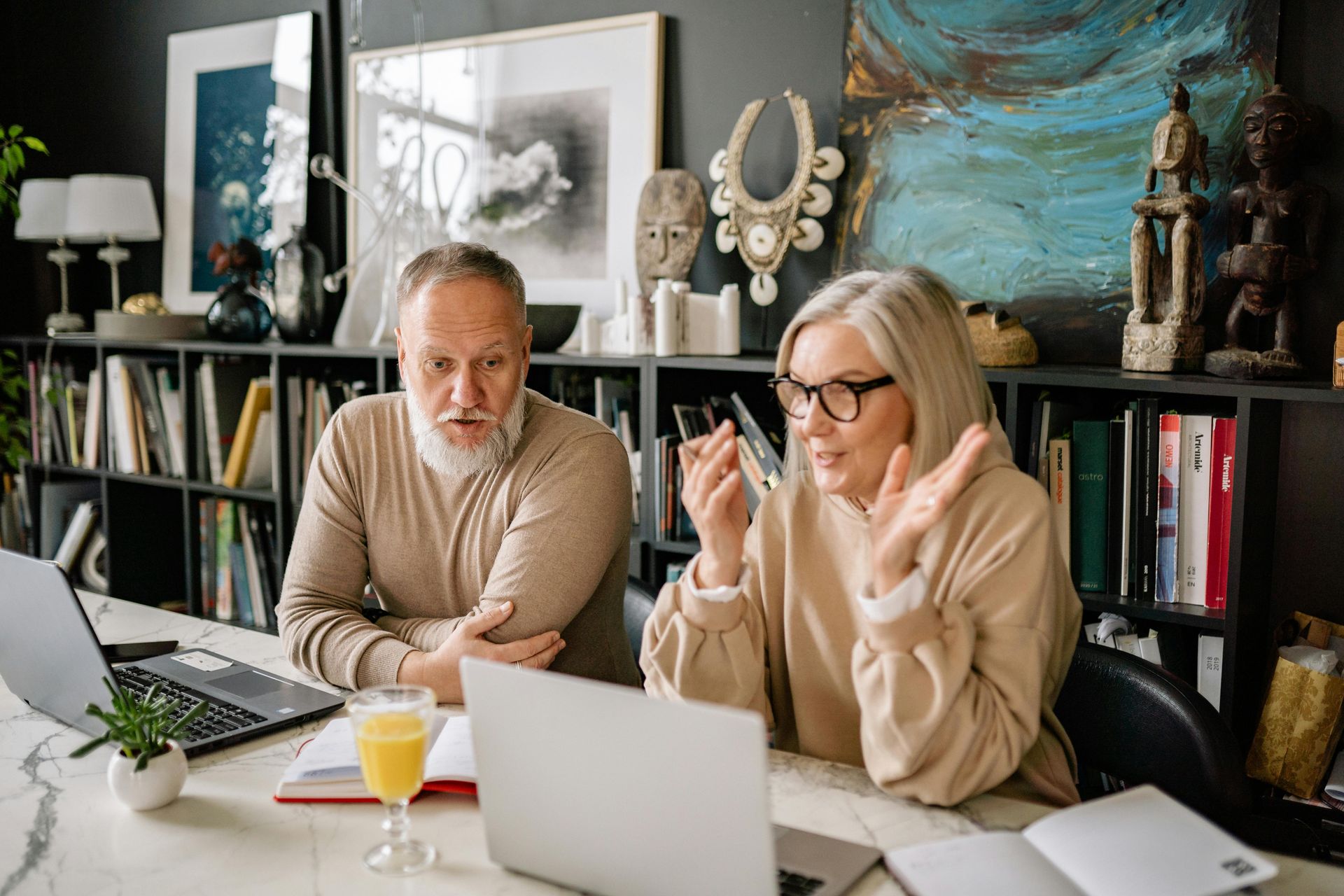 Senior couple at a desk with laptops, discussing. Woman gestures, man looks on. Bookshelves in background.