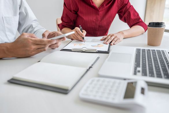 Two people at a desk reviewing documents, with a laptop, calculator, and coffee.