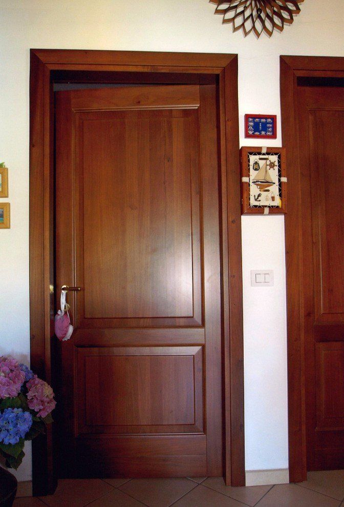 Porta in legno con telaio scuro, maniglia e due pannelli. Fiori viola e rosa a sinistra, opere d'arte e interruttore della luce a destra.
