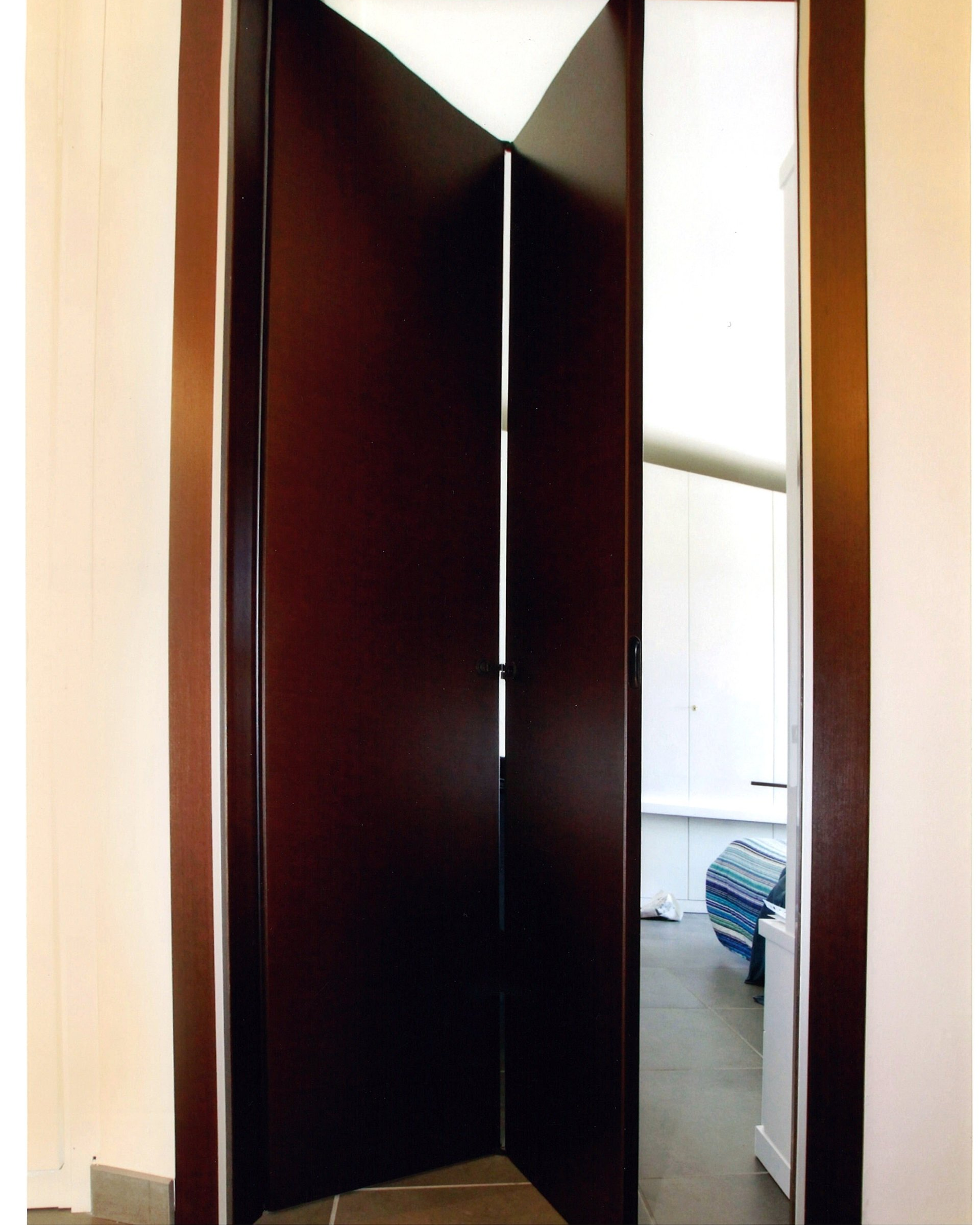 Porta a soffietto marrone scuro, parzialmente aperta, in un corridoio dalle pareti bianche. Vista su una stanza arredata.