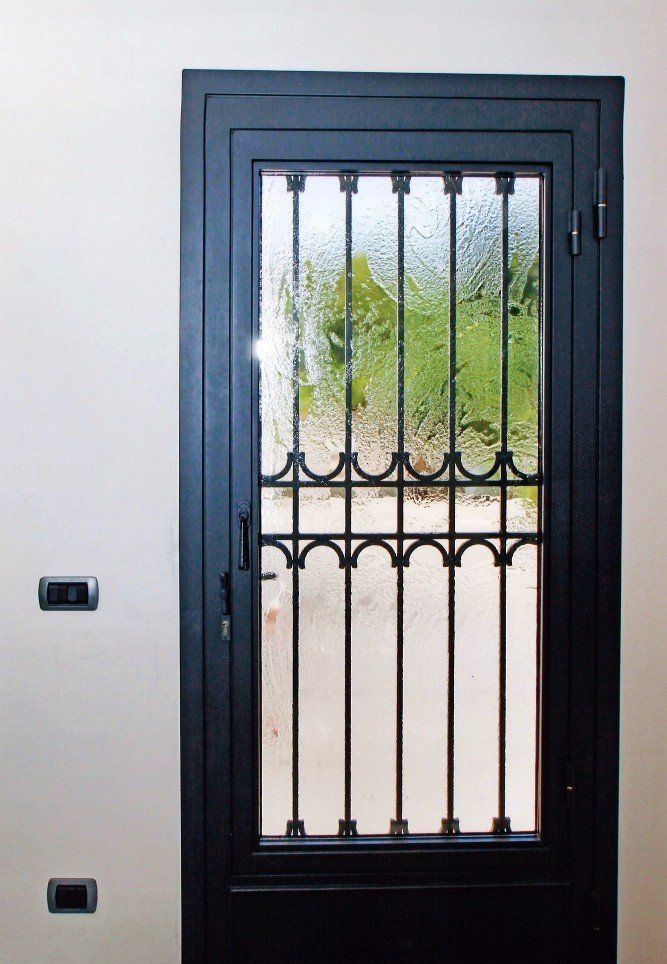 Porta in metallo nero con pannello in vetro e barre decorate contro una parete bianca.