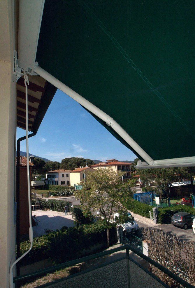 Una tenda verde si estende su un balcone, con vista su una strada con edifici e alberi sotto un cielo azzurro.