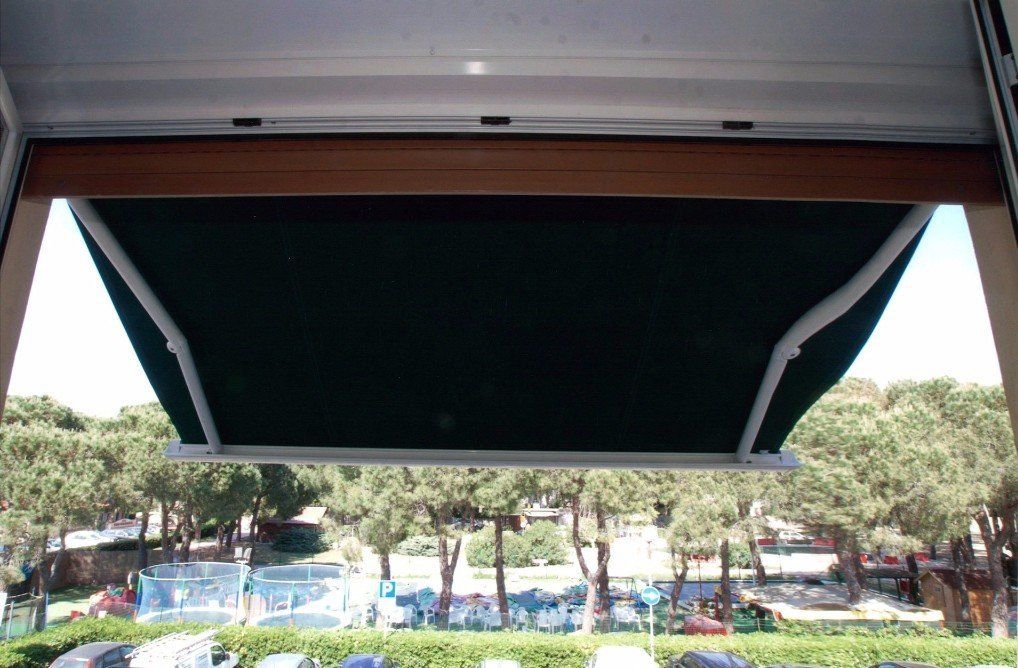 Una tenda da sole verde scuro parzialmente estesa sopra una finestra, da cui si vedono alberi e cielo all'esterno.