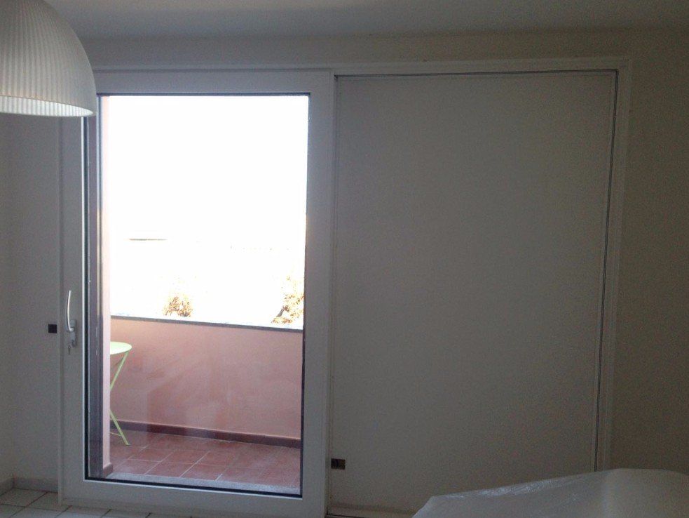Porta scorrevole in vetro che si apre su un balcone, con telaio bianco. Porta scorrevole bianca chiusa sulla destra.
