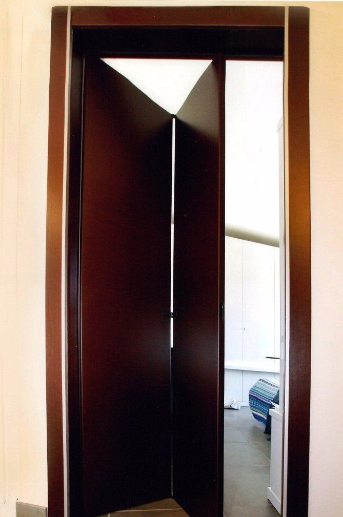 Porta girevole in legno scuro, aperta su una stanza con parete bianca, luce naturale e mobili.