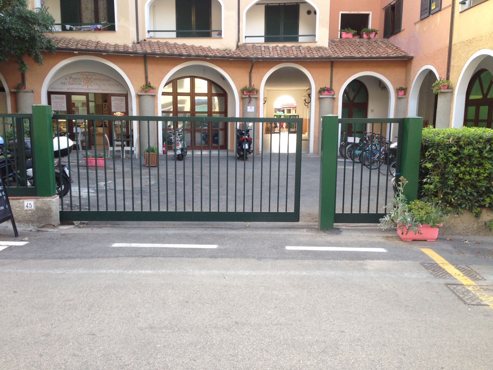 Cancello metallico verde che si apre su un edificio con porte ad arco. Motociclette e biciclette visibili.