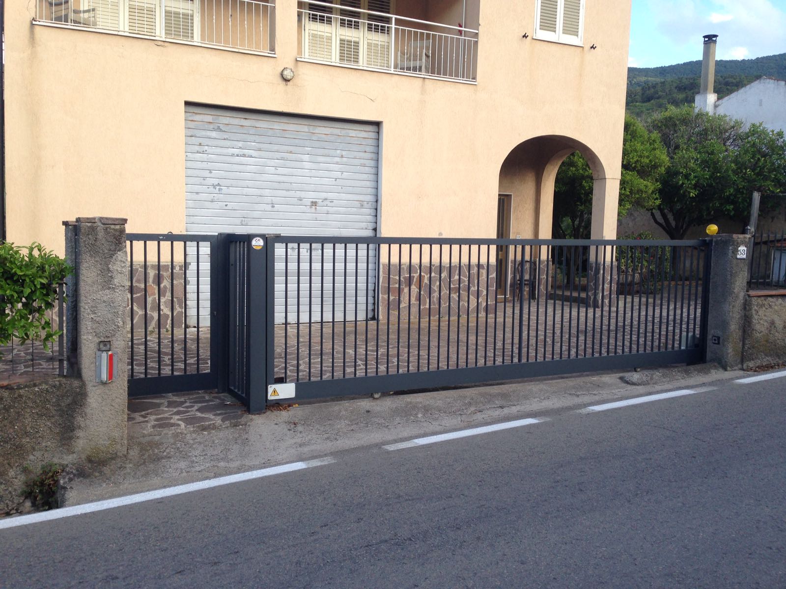 Cancello scorrevole in metallo grigio davanti a un edificio a due piani con porta da garage e ingresso ad arco.