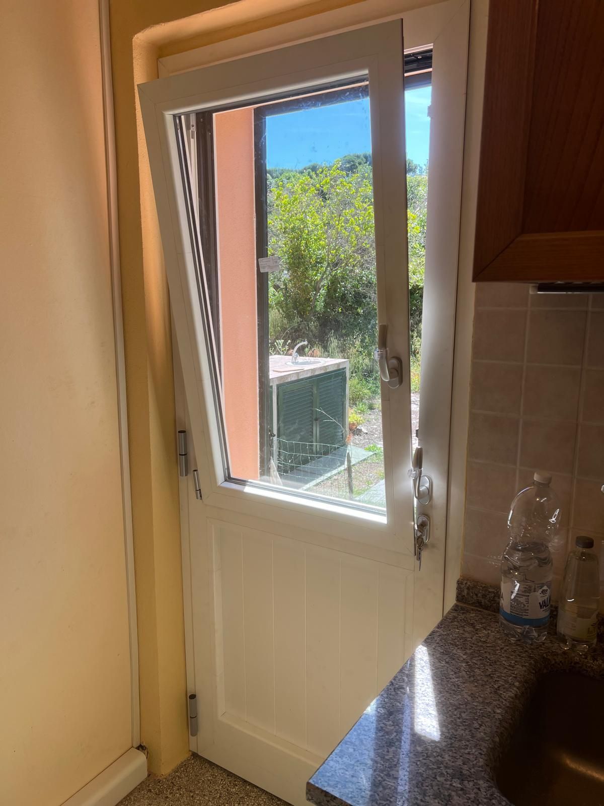 La porta bianca della cucina si apre su una soleggiata vista esterna. Gli interni sono caratterizzati da un piano di lavoro in granito e pareti beige.