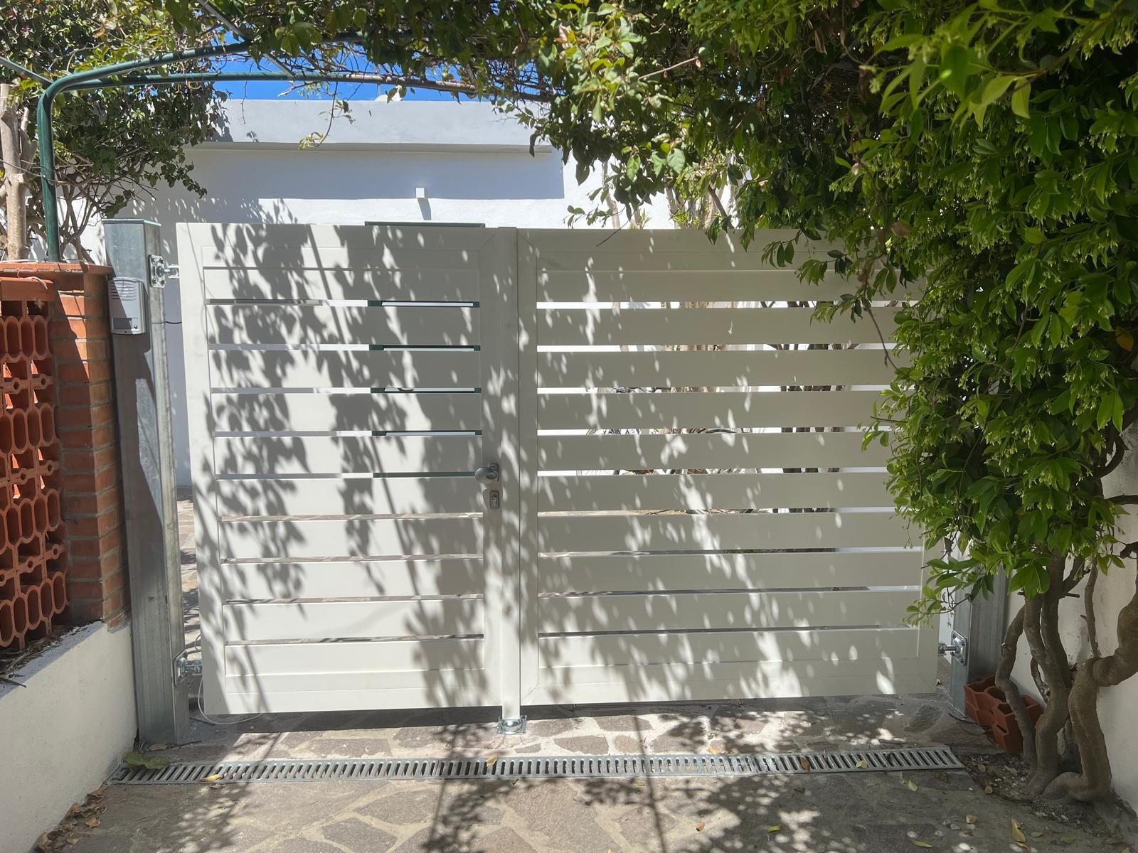 Cancello bianco a stecche orizzontali davanti a un edificio, parzialmente ombreggiato dagli alberi.