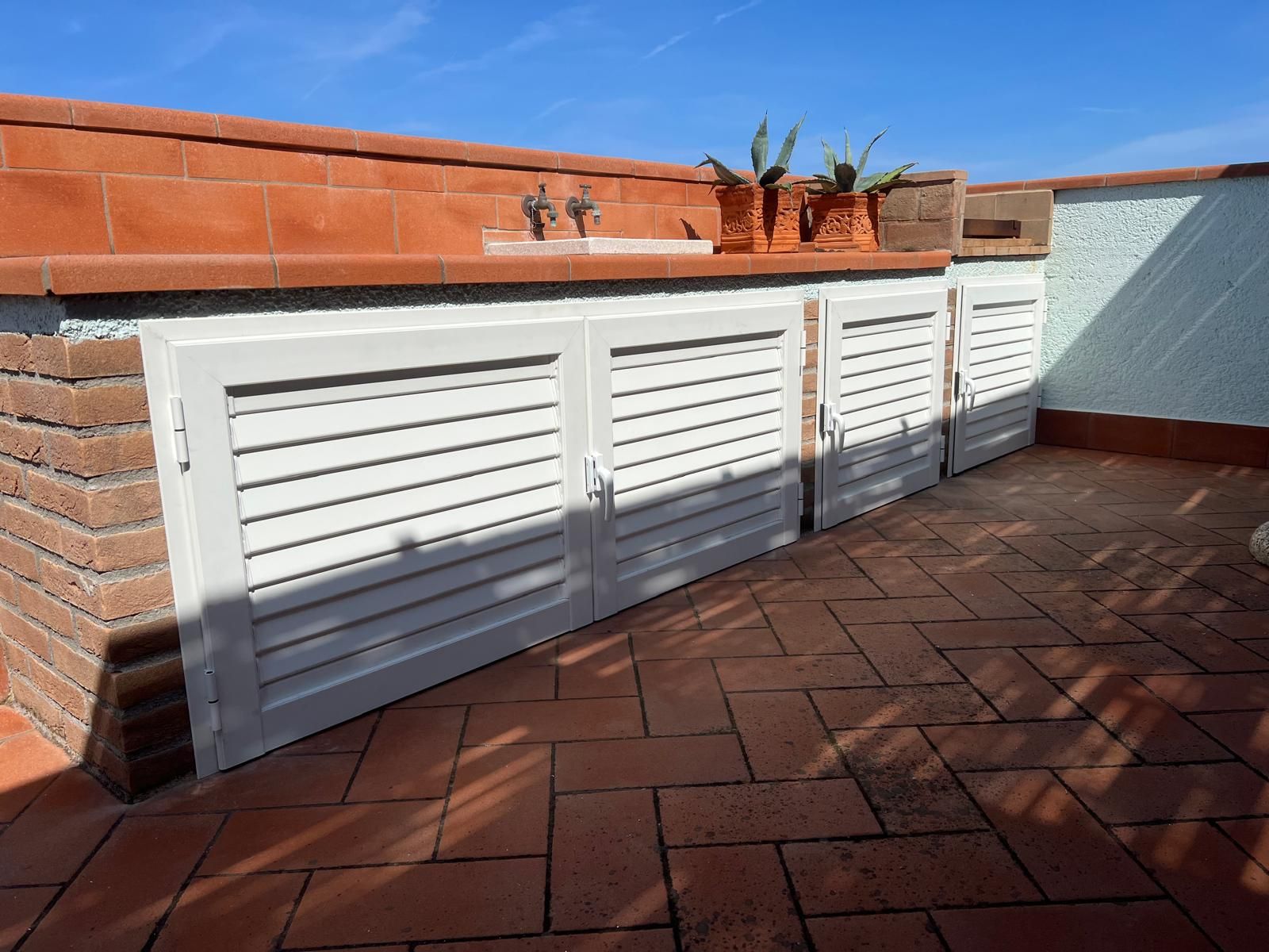 Cucina esterna con piastrelle in terracotta, ante dei mobili con persiane bianche, paraschizzi in mattoni e rubinetti dell'acqua, sotto il cielo azzurro.