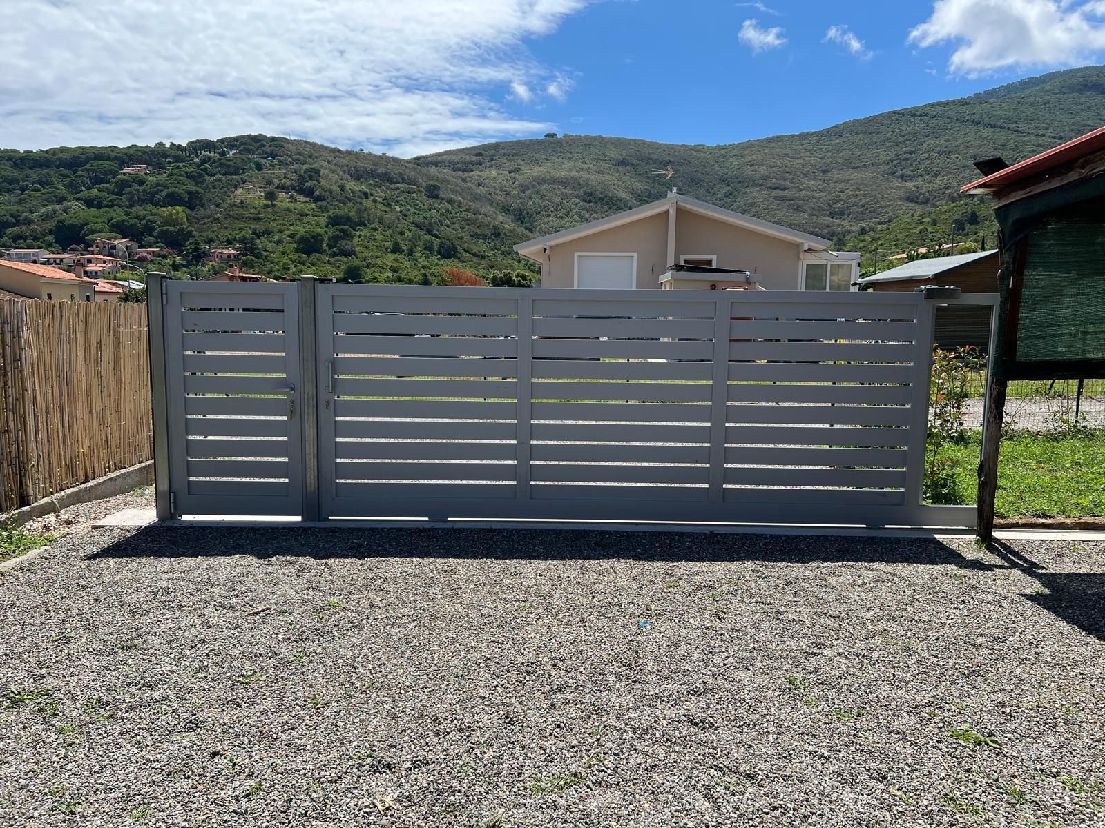 Cancello orizzontale grigio con stecche davanti a un vialetto di ghiaia, una casa e montagne sullo sfondo.