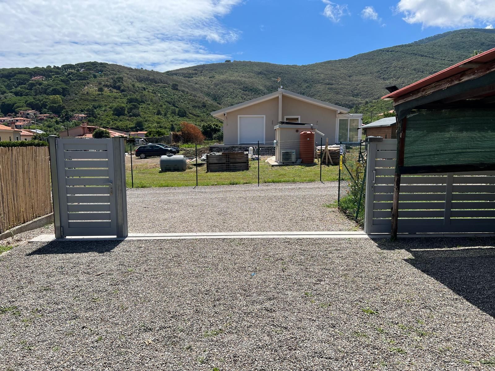 Vialetto di ghiaia con cancelli grigi aperti che conduce a una casa con le montagne sullo sfondo in una giornata di sole.