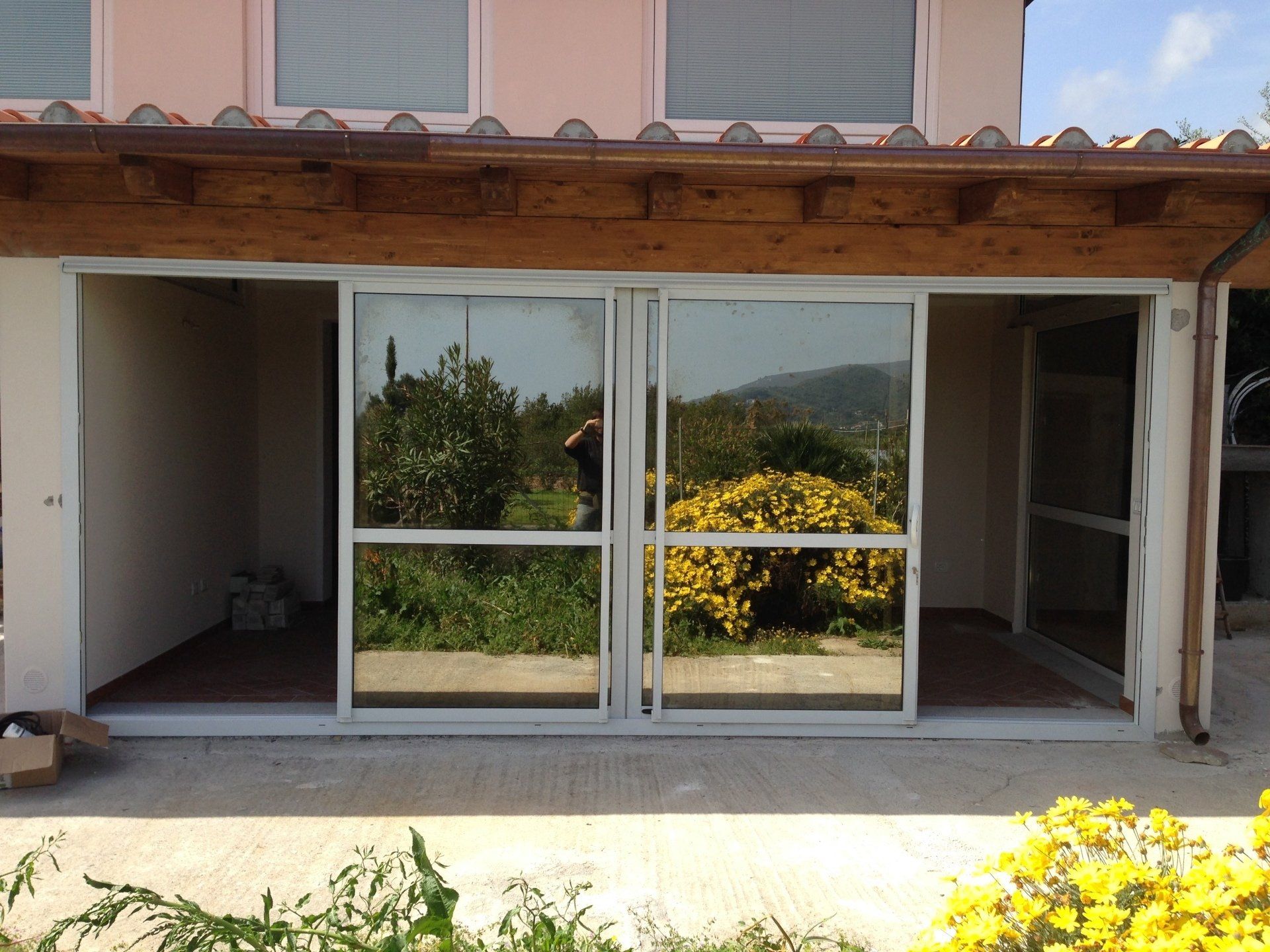 Porte scorrevoli in vetro che si aprono su uno spazio esterno soleggiato. Infissi bianchi, finestre riflettenti, vegetazione a vista.