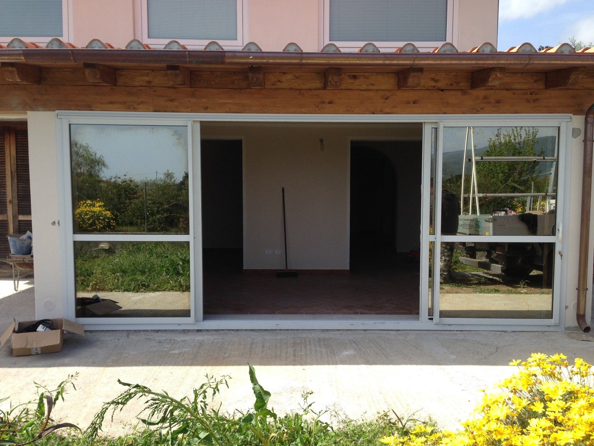 Vista esterna di un portico con porte scorrevoli in vetro, pavimento in cemento e fiori gialli.