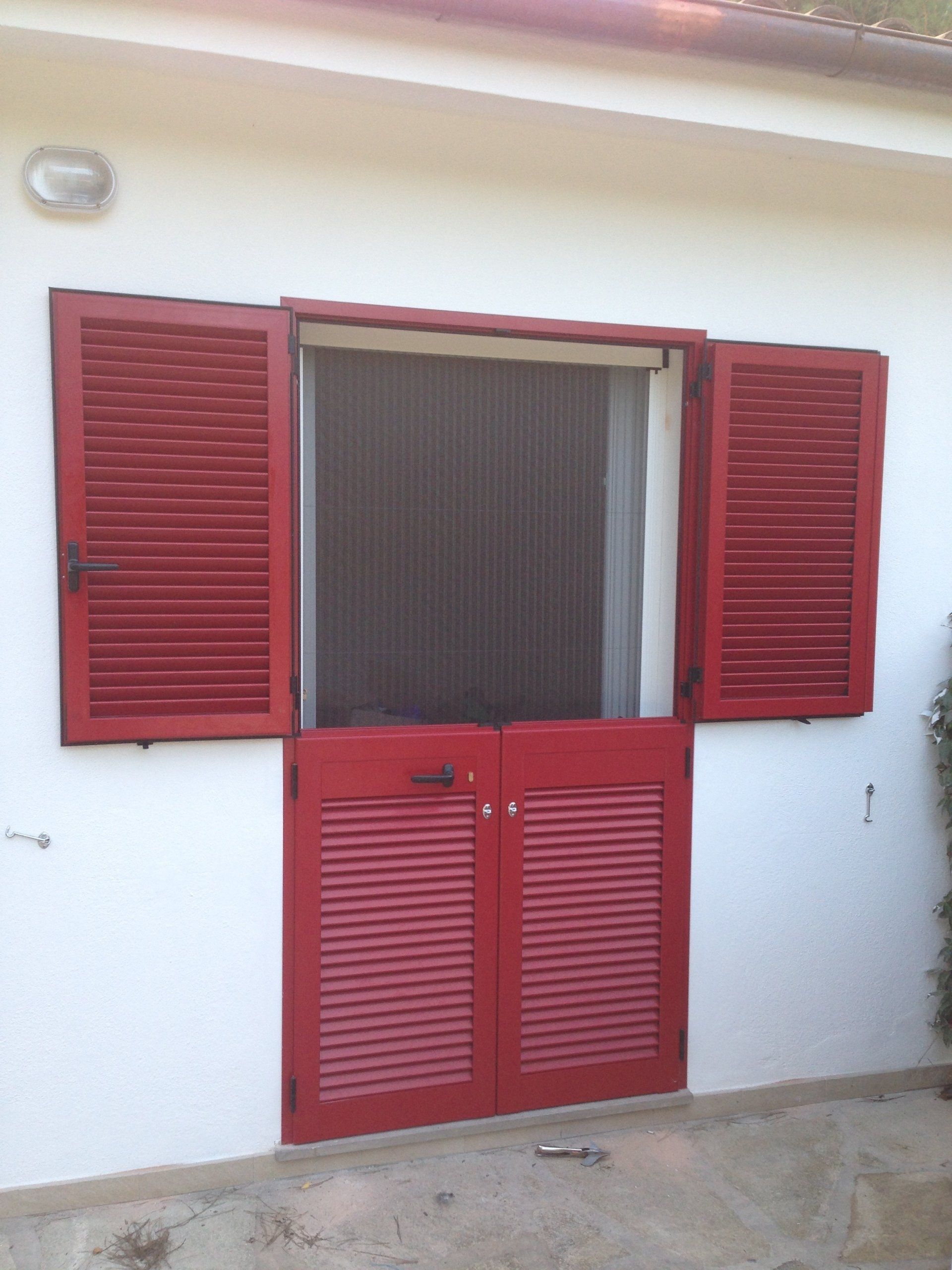 Persiane e porta rosse su un edificio bianco. Le persiane sono aperte per rivelare una zanzariera.