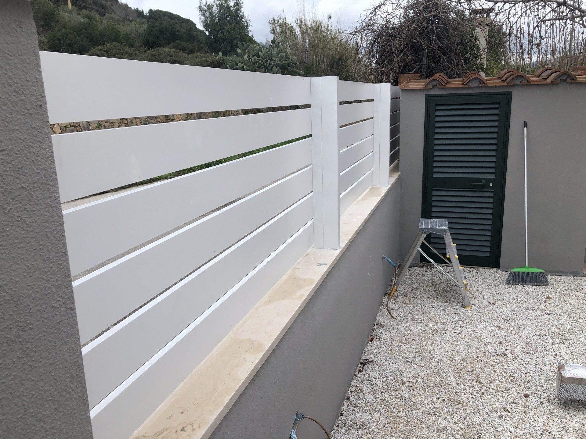 Recinzione grigia con stecche orizzontali in cima a un muretto basso, accanto a una piccola porta e ghiaia.