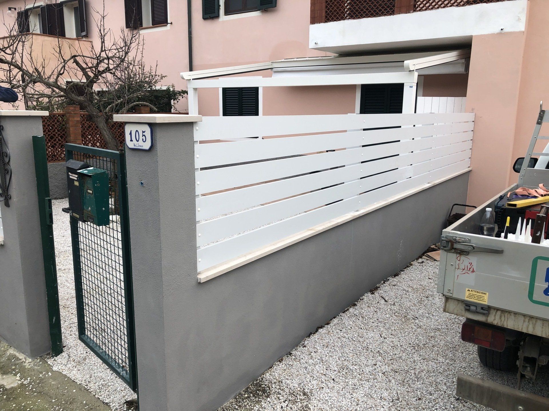 Recinzione bianca con doghe orizzontali, base in cemento grigio, piccolo cancello verde ed edificio rosa.