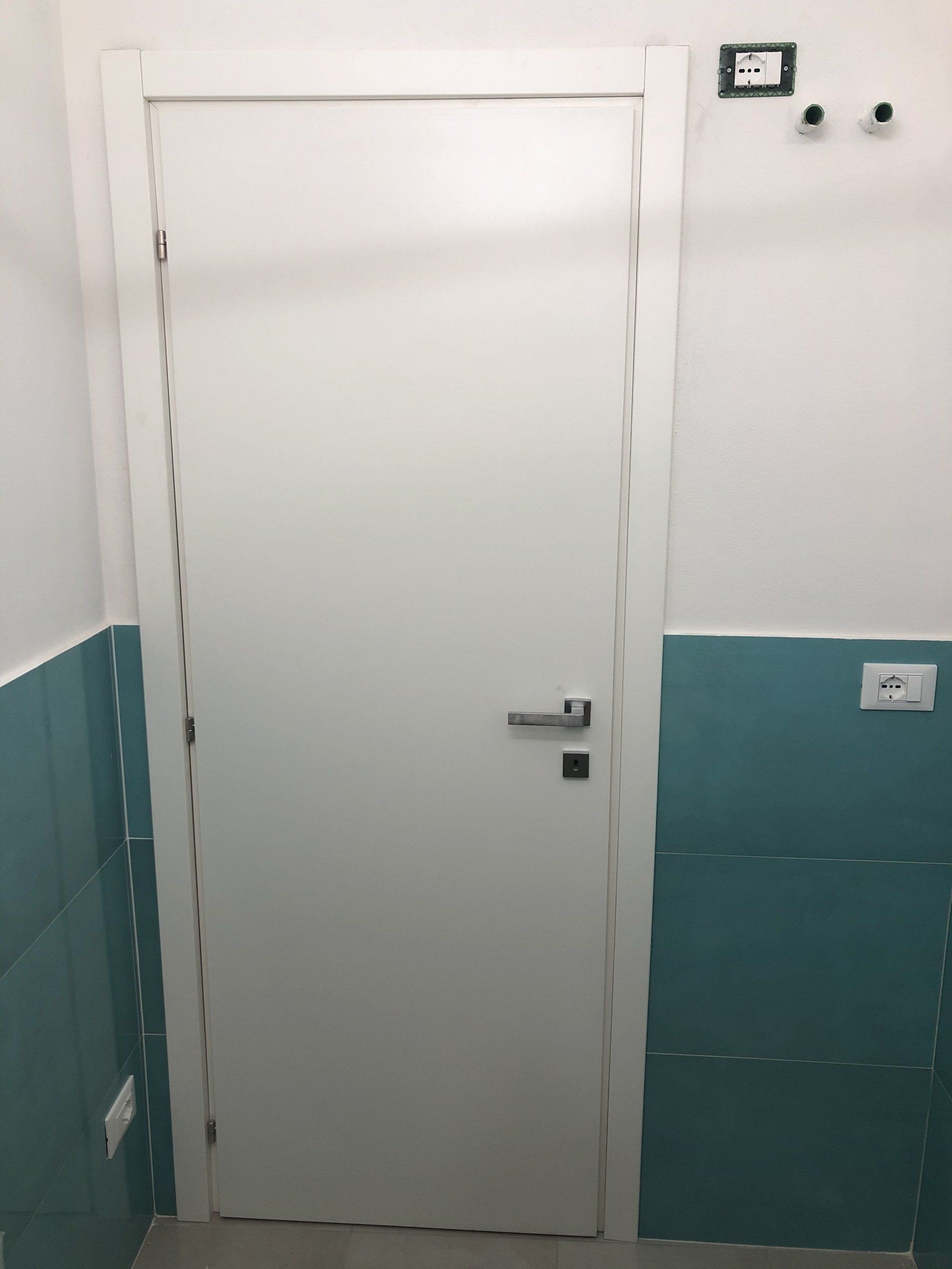 Porta bianca in una stanza con piastrelle color verde acqua e finiture bianche, prese elettriche e ganci appendiabiti.